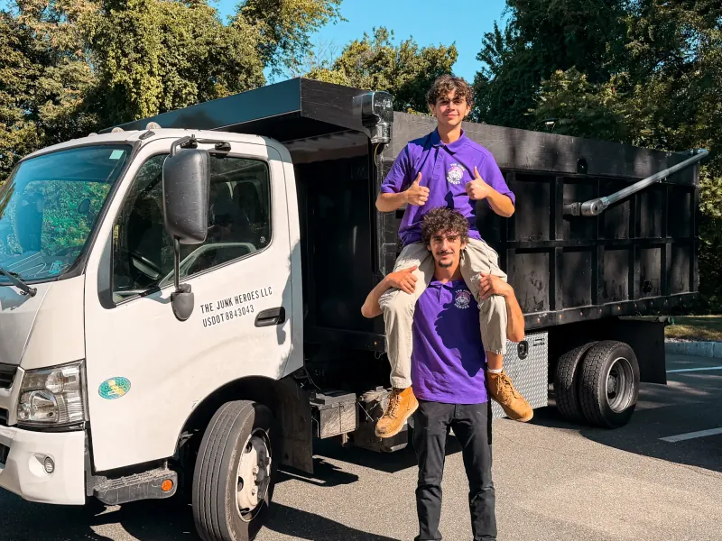 Henrique and Filipe of The Junk Heroes in branded uniforms, giving a thumbs up in front of their junk removal dump truck on the North Shore, MA
