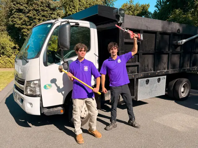 Henrique and Filipe of The Junk Heroes holding a sledgehammer and a reciprocating saw beside their dump truck, ready for a light demolition job in Marblehead, MA